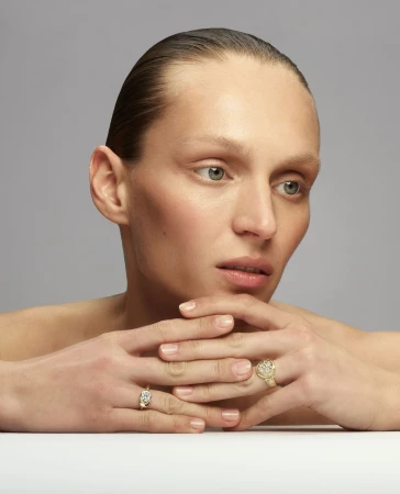 Close-up studio shot of a model with slicked-back hair wearing gold rings with neutral makeup, resting chin on interlaced fingers.