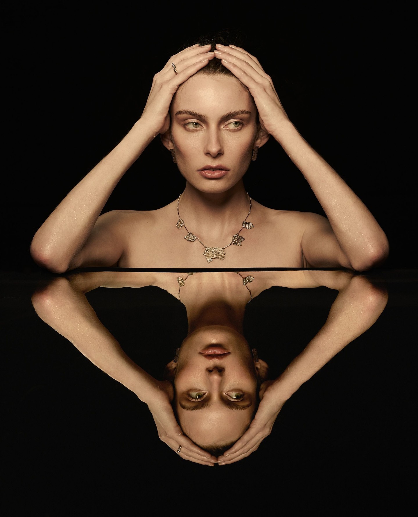 Model with hands on head wearing a gold necklace, reflected on a dark mirrored surface against a black background.