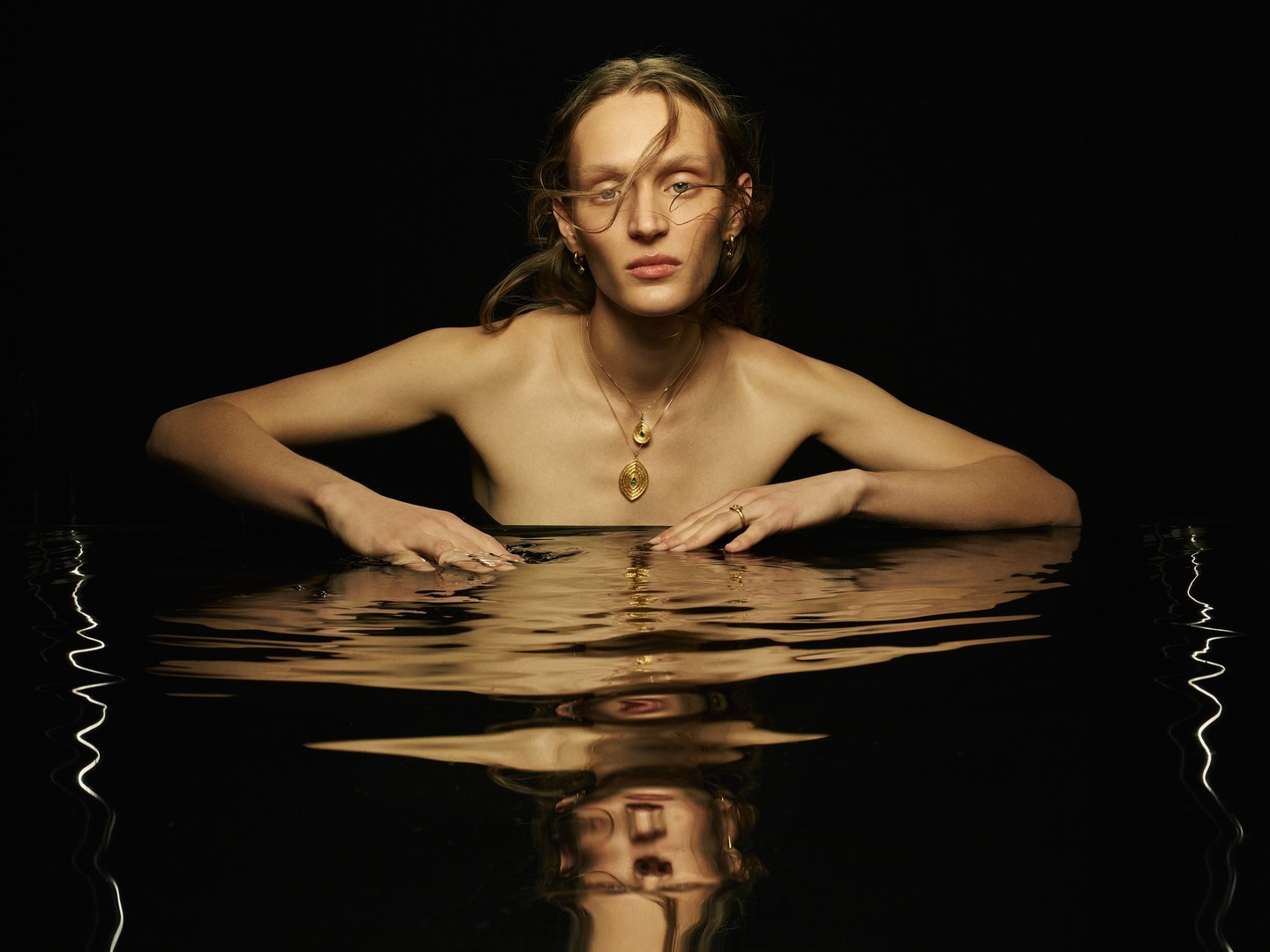 Portrait of an adult female partially submerged in water against a dark background, wearing gold jewelry.