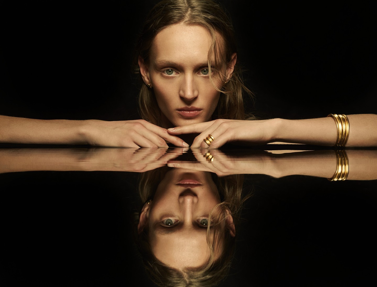 Elegant portrait of a blonde woman reflecting on a black surface, wearing gold bangles and rings, against a dark background.