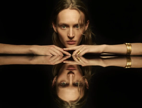 Elegant portrait of a blonde woman reflecting on a black surface, wearing gold bangles and rings, against a dark background.