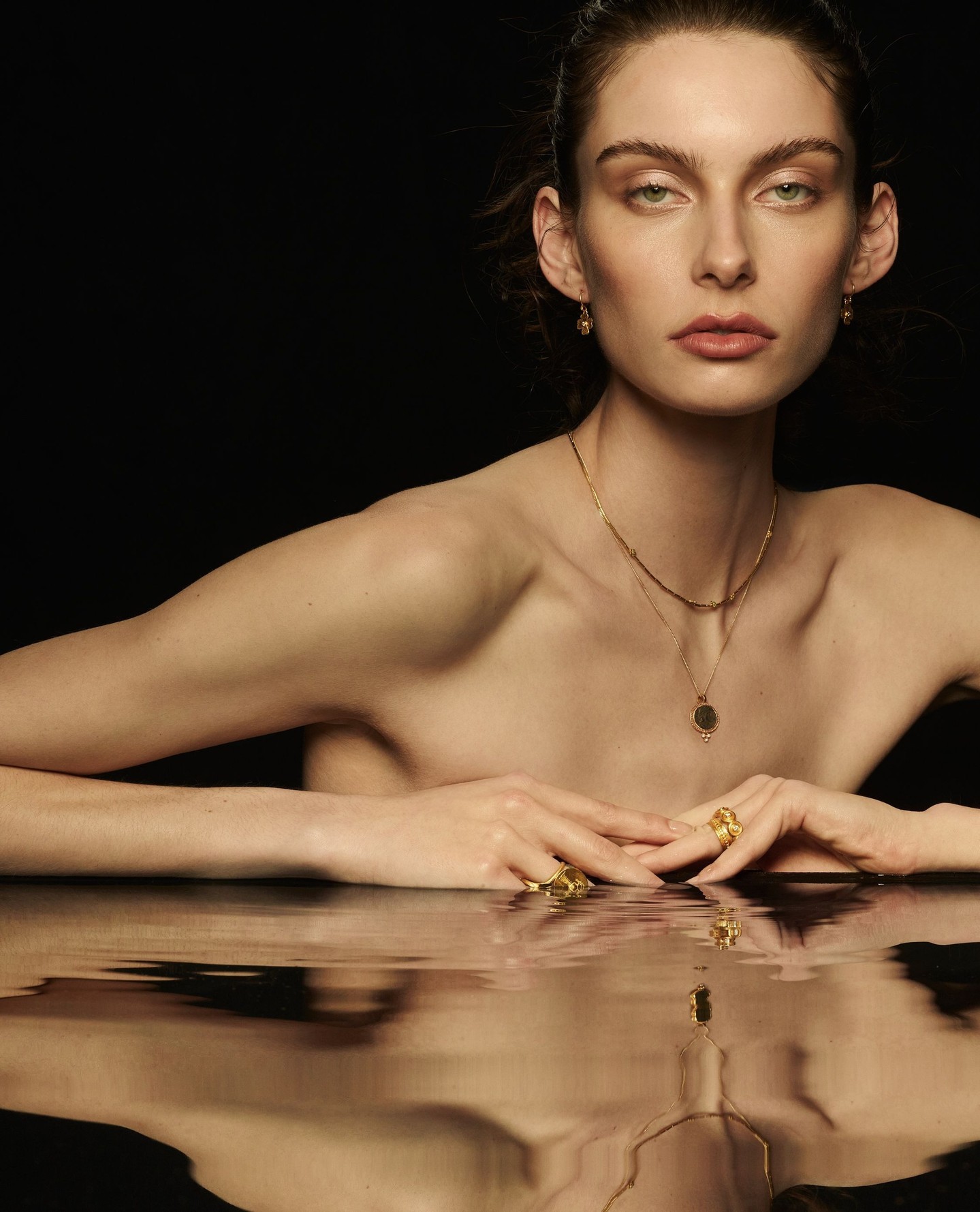 Model wearing delicate gold necklaces and rings, reflected in water against a dark background for Temple of the Sun.