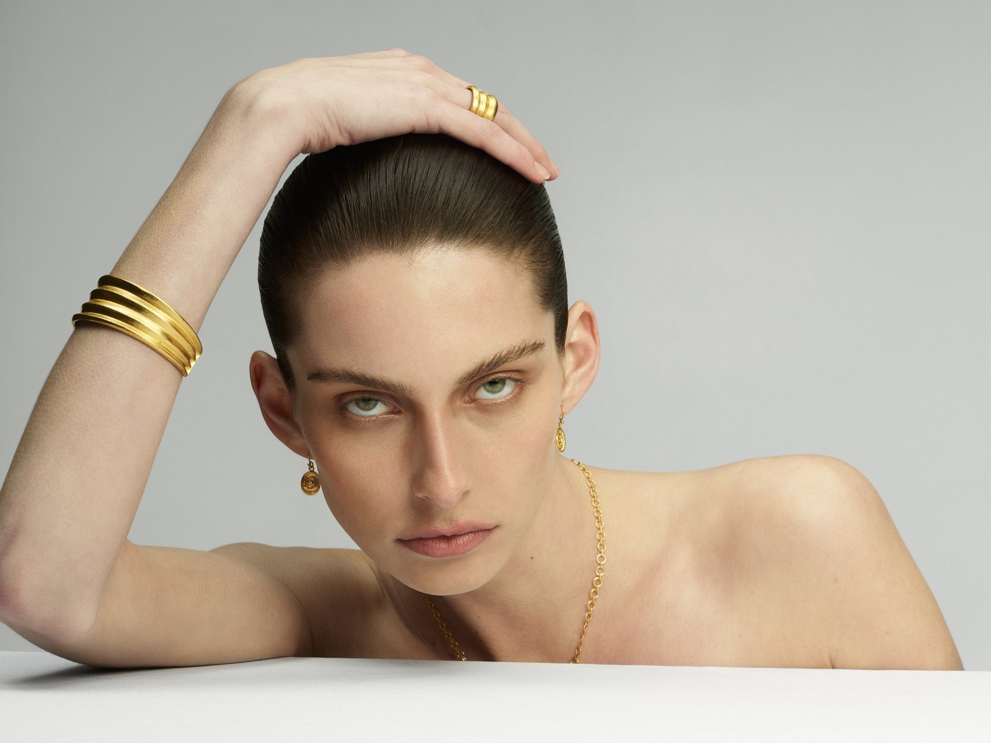 Close-up shot of a female model wearing gold jewelry, including a ring, a bracelet, a necklace, and earrings, against a neutral backdrop.