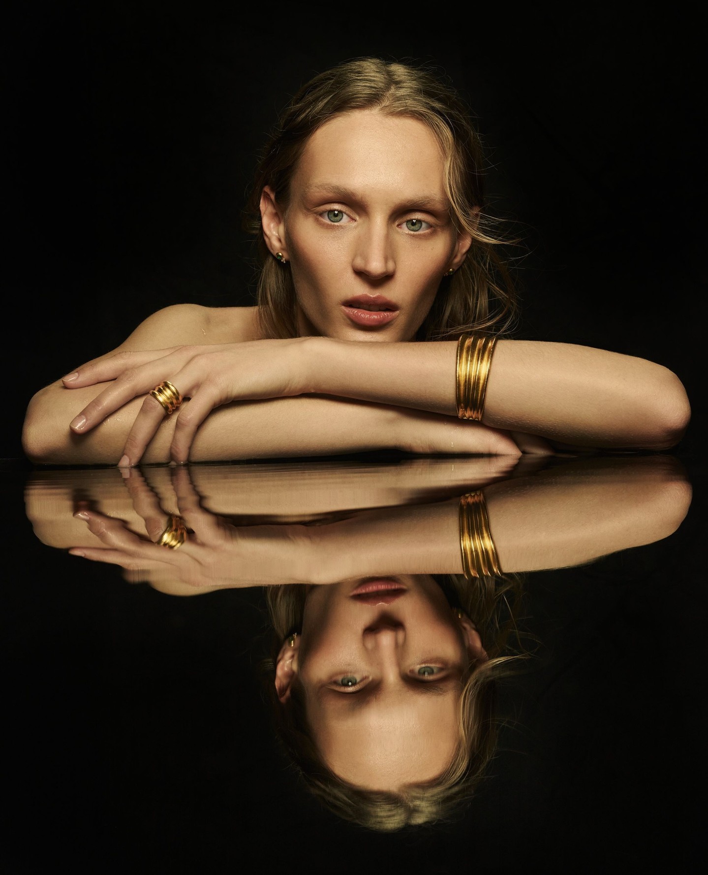 Portrait of a blonde woman with gold jewelry including multiple bracelets and rings, reflected on a glossy black surface.
