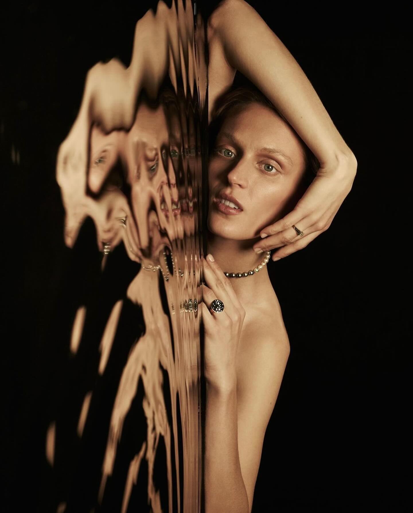 Striking beauty shot of a fair-skinned adult woman with pearl necklace and rings posing in front of a dark background and wavy glass.