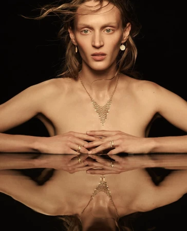 Model wearing gold necklace and earrings with hands reflected on a dark glossy surface against a black background.