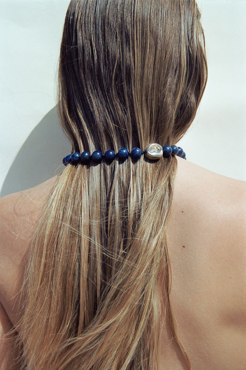 View from behind of a woman with long blonde hair, wearing a blue beaded necklace with a silver bead on a white background.