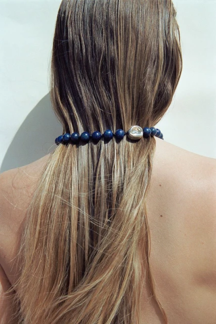 View from behind of a woman with long blonde hair, wearing a blue beaded necklace with a silver bead on a white background.