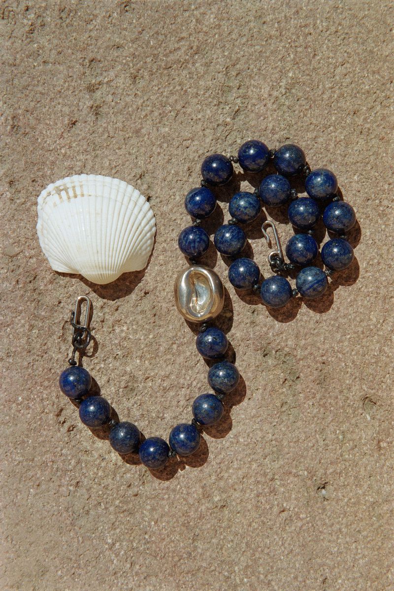 Still life of a beaded necklace with blue lapis lazuli beads, a silver accent bead, and a white seashell on a textured tan background.