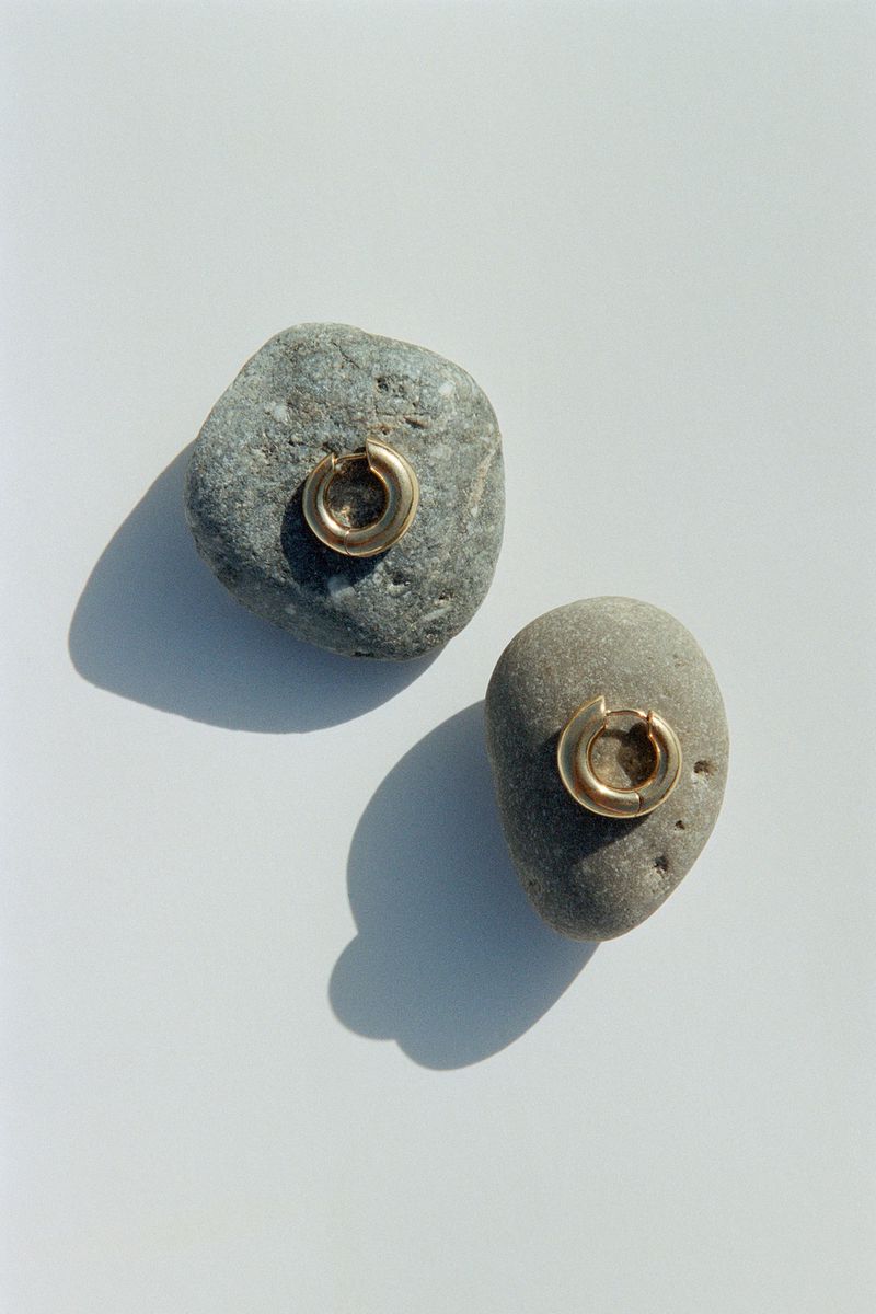Two gold hoop earrings displayed atop small grey rocks on a bright white surface, casting soft shadows, jewelry product shot.