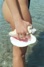 Close-up of a person holding a large white conch shell standing in shallow clear ocean water, shot for Studio Deve.