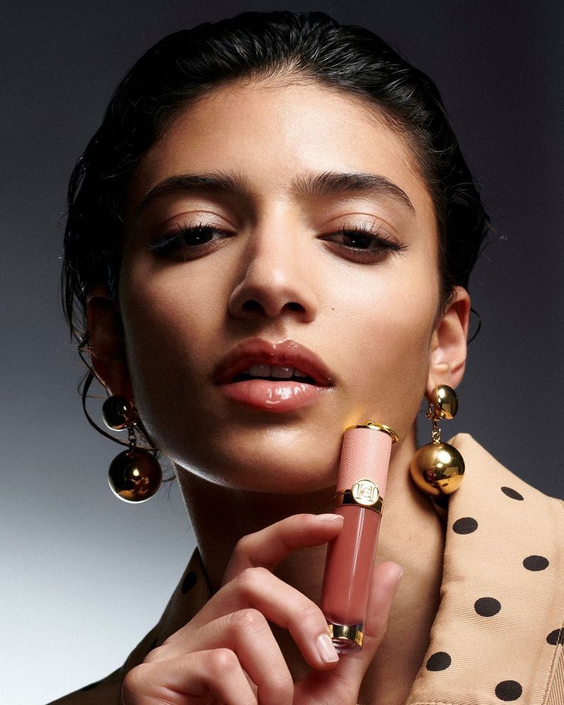 Close-up shot of a woman with dark hair posing while holding a tube of pink lip gloss, wearing gold earrings and a tan polka-dot jacket.