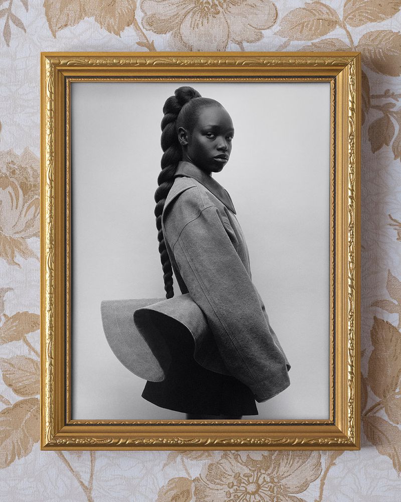 Monochromatic portrait of a Black woman with braided hair, wearing a gray dress, presented in a gold frame against floral wallpaper.
