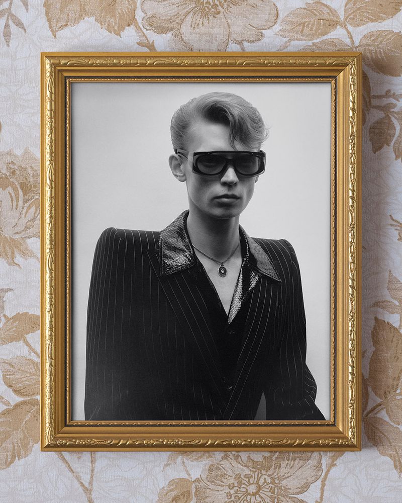 Monochrome studio portrait of androgynous person with retro hairstyle and sunglasses, wearing pinstripe suit, displayed in a gold picture frame.