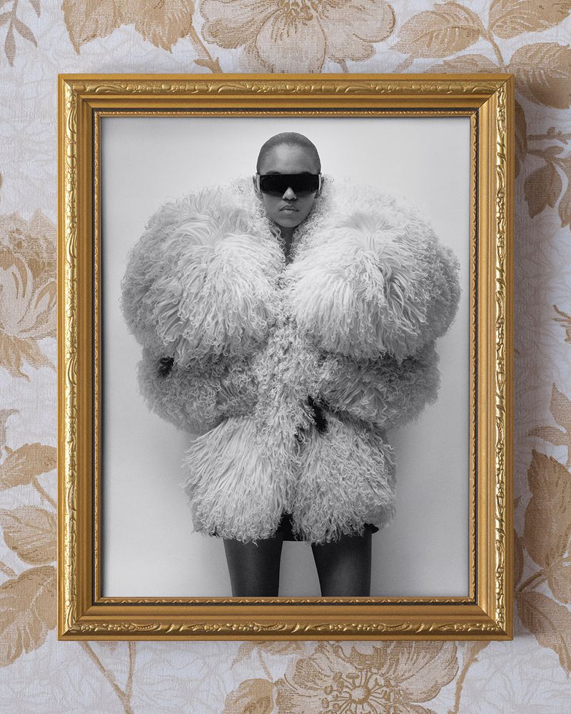 Fashionable adult in an oversized fluffy coat and sunglasses, presented in a gold frame against a floral wallpaper background.
