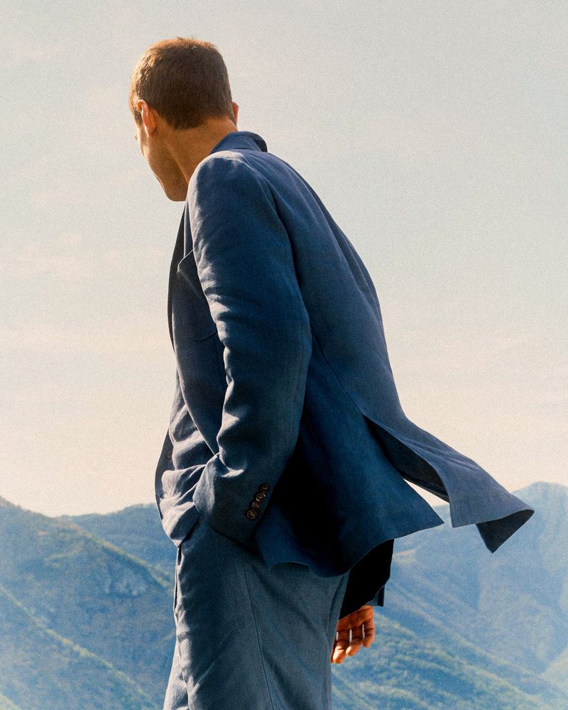 A man in a blue suit stands on a hilltop with mountains in the background, back to the camera, looking down during daytime.