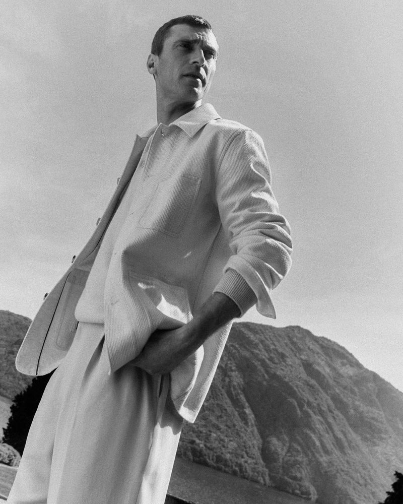Monochrome portrait of a man in a white linen suit posing outdoors against a backdrop of rolling hills and a clear sky.