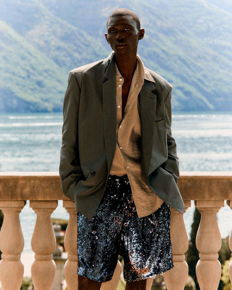 A stylish African-American man models olive blazer with unbuttoned tan shirt and sequined shorts outdoors against a lake and mountain view.