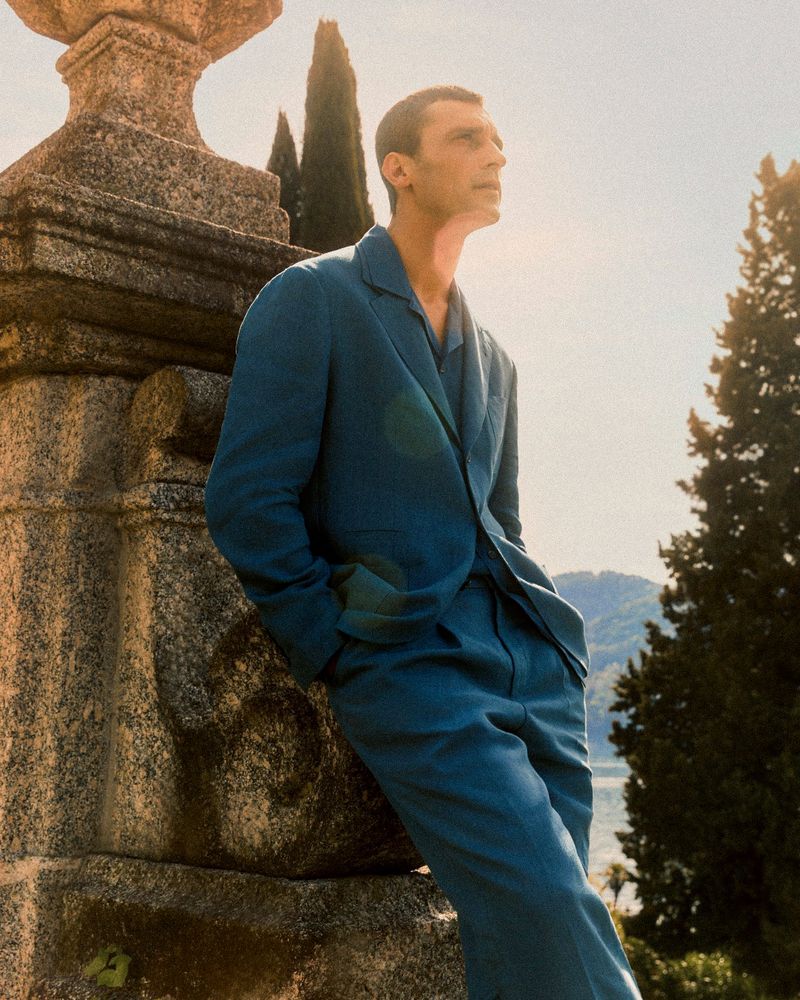 Stylish adult man in a teal blue suit leans against a stone pillar outdoors with trees and lake view in the background on a sunny day.