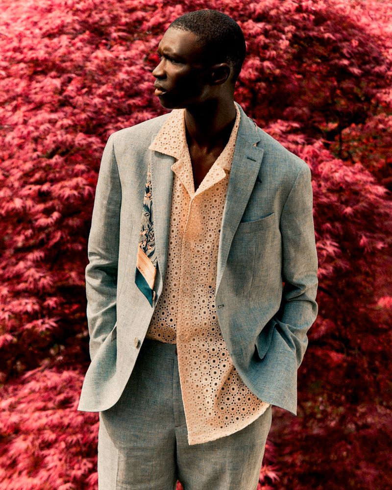 Dark-skinned male model in a light blue suit poses outdoors against a reddish-pink foliage backdrop. Fashion editorial style, medium shot.