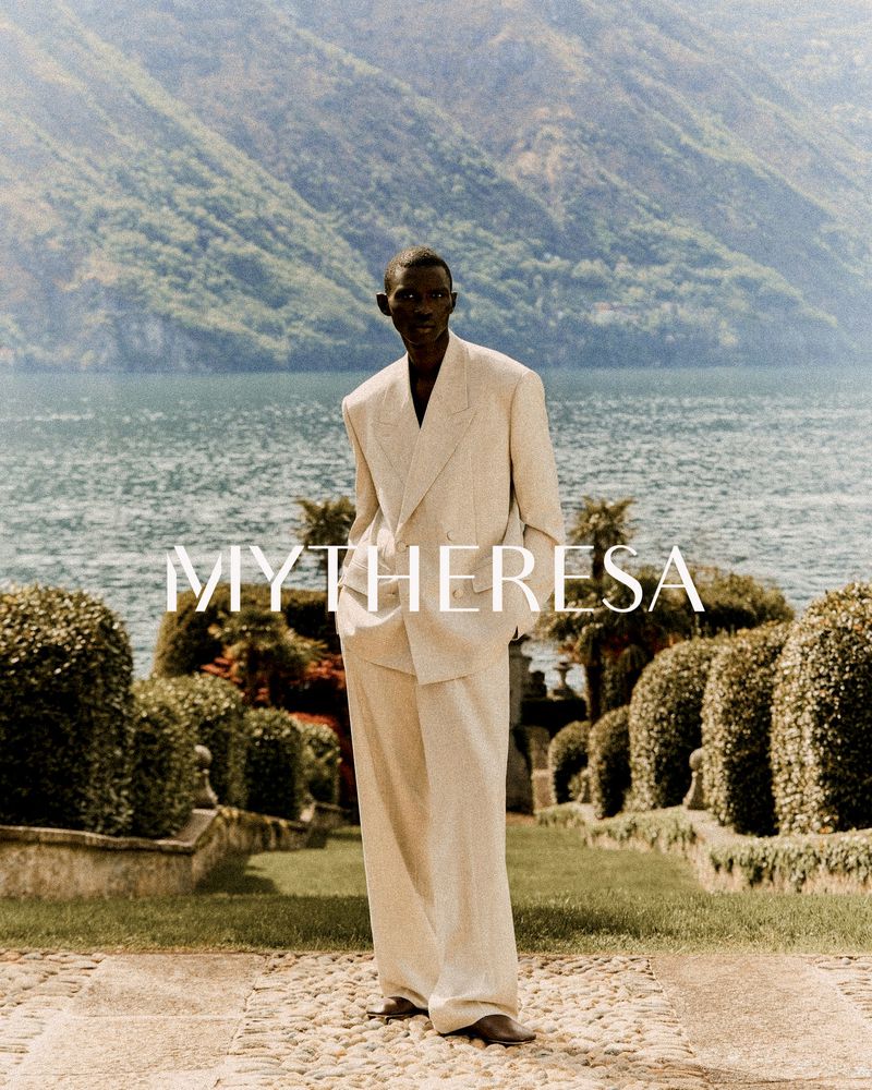 Elegant, dark-skinned man in cream-colored suit stands on stone path against backdrop of Lake Como and lush, green mountains.