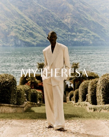 Elegant, dark-skinned man in cream-colored suit stands on stone path against backdrop of Lake Como and lush, green mountains.