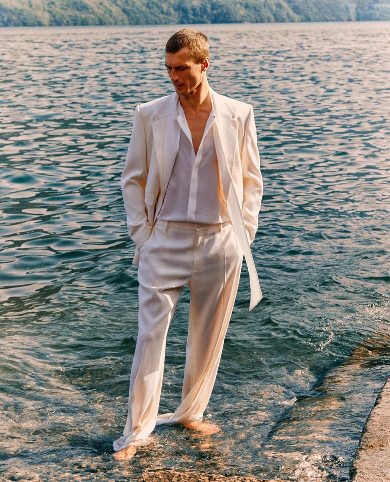 Man in a white suit standing in water near a lake with mountains in the background, gazing downward with a contemplative expression.
