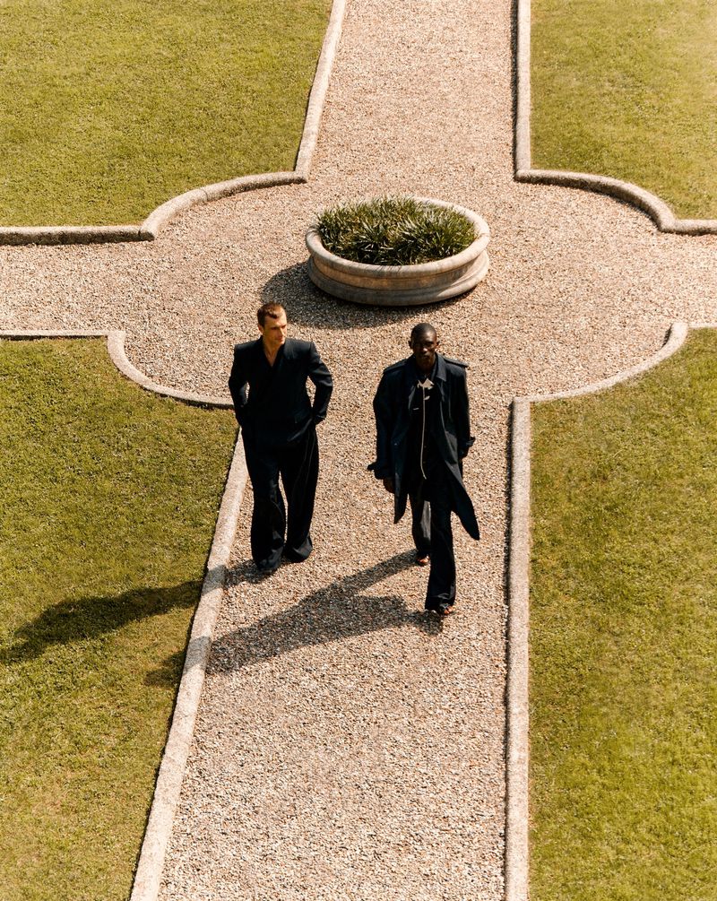 Two men in dark suits walk on a stone path through a manicured lawn and landscaping, captured from a high angle.