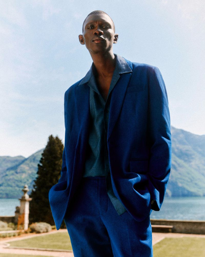 A man poses in a blue suit outdoors with mountains and water in the background, standing on a manicured lawn in natural light.