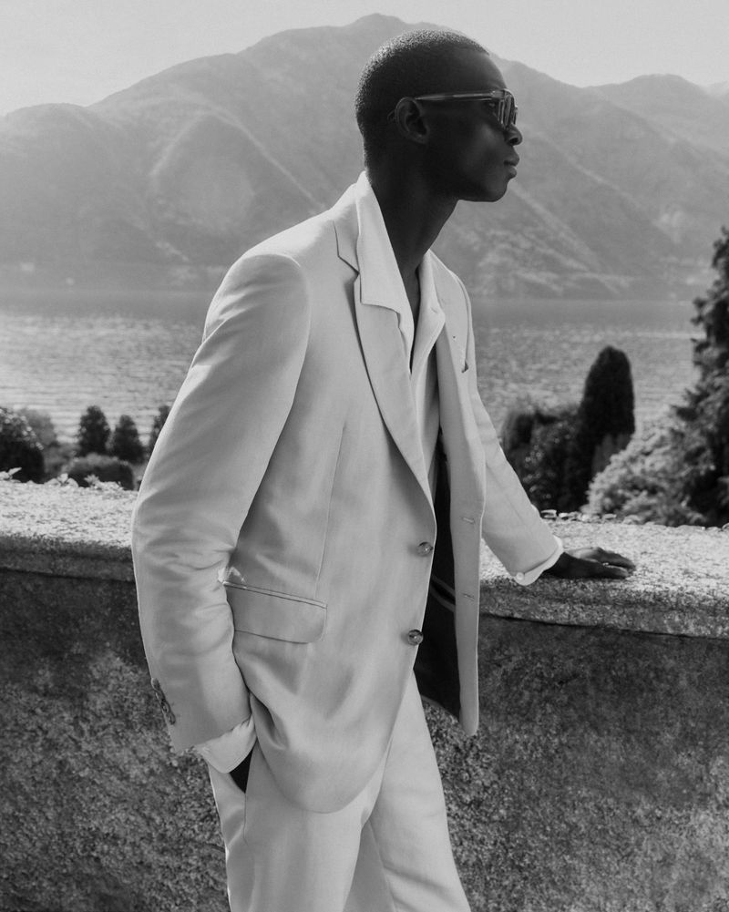 Black male model in a light suit and sunglasses poses on a stone ledge against a mountain and lake backdrop in black and white.