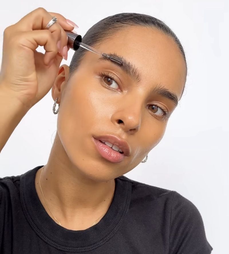 A woman with fair skin applies brow serum to her eyebrows with a dropper in a beauty shot against a white backdrop. 