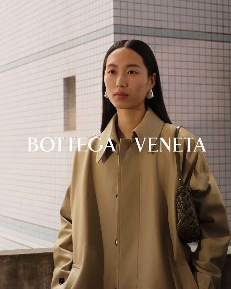 Asian model presents Bottega Veneta beige trench coat and green woven purse in a studio shot with a tiled wall background.