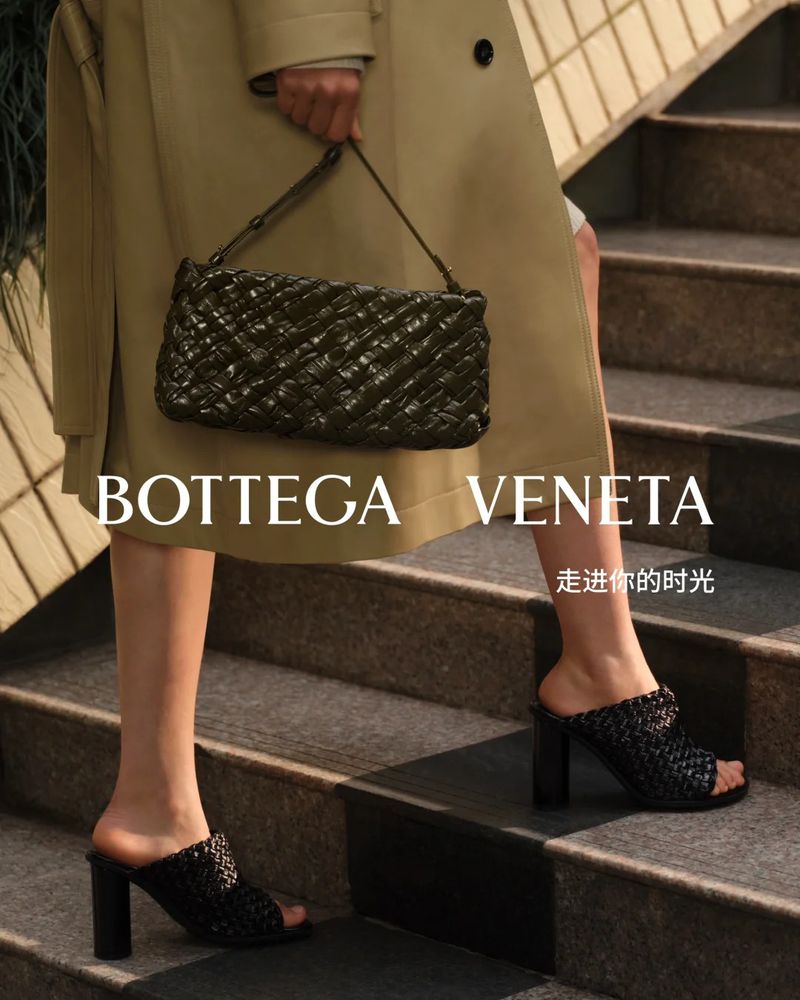Close-up of a fashion model wearing a tan coat and black woven mules on stairs, carrying a dark green woven Bottega Veneta handbag.