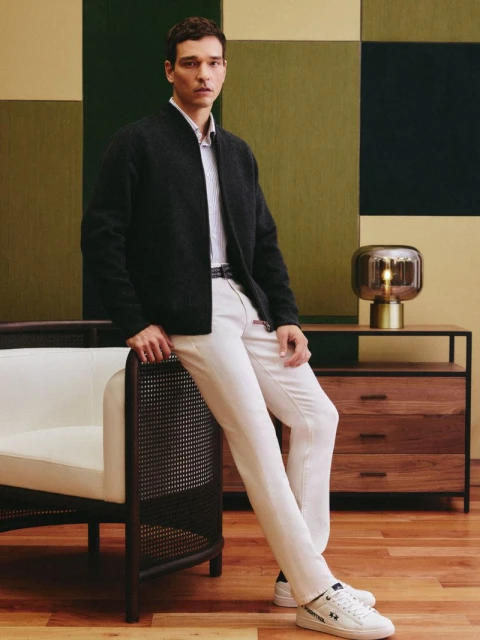 Fashionable adult male in a black jacket and white trousers poses indoors near a mid-century style table lamp and geometric wall.