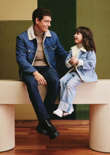 A father and daughter are sitting on a bench, smiling at each other while wearing matching denim jackets with beige turtlenecks.