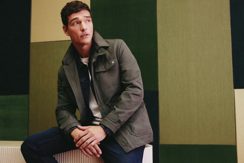 Portrait of a handsome adult man sitting and posing in a grey jacket and blue jeans against a geometric wall background.