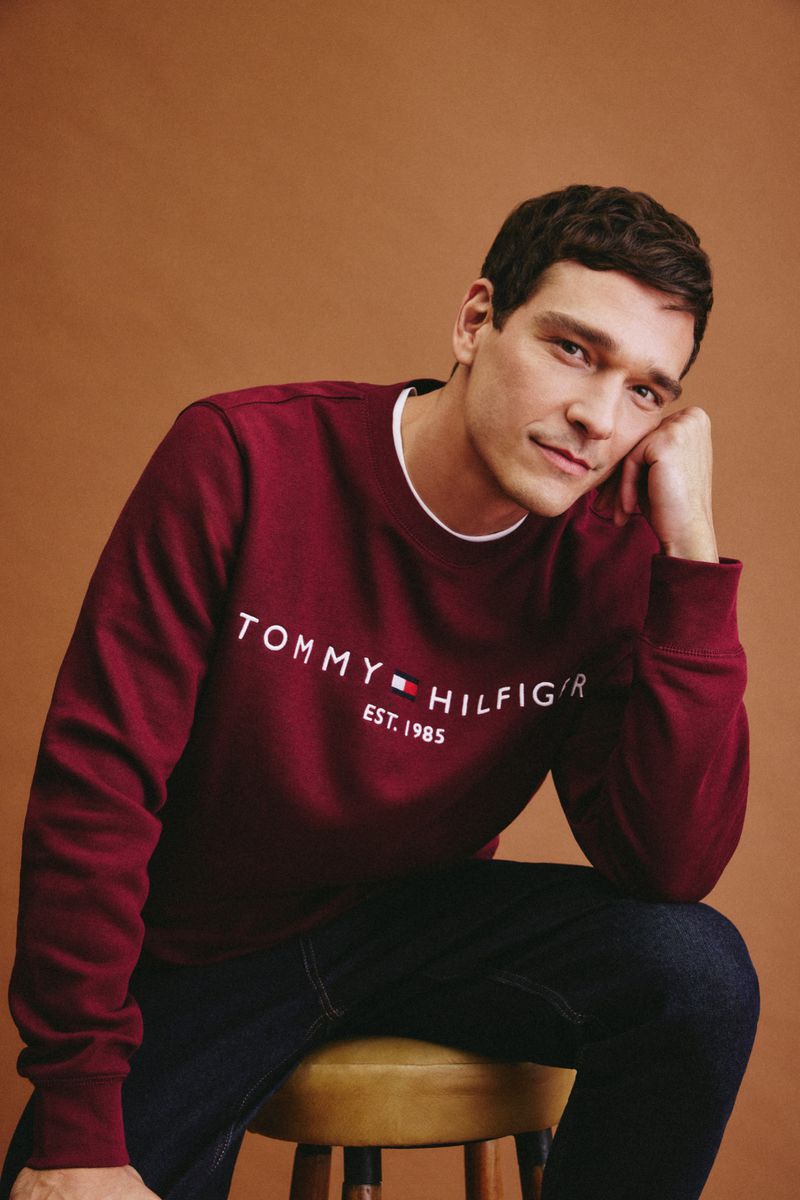 A brunette adult male sits on a stool wearing a Tommy Hilfiger sweatshirt and jeans against a brown background in a studio shot.