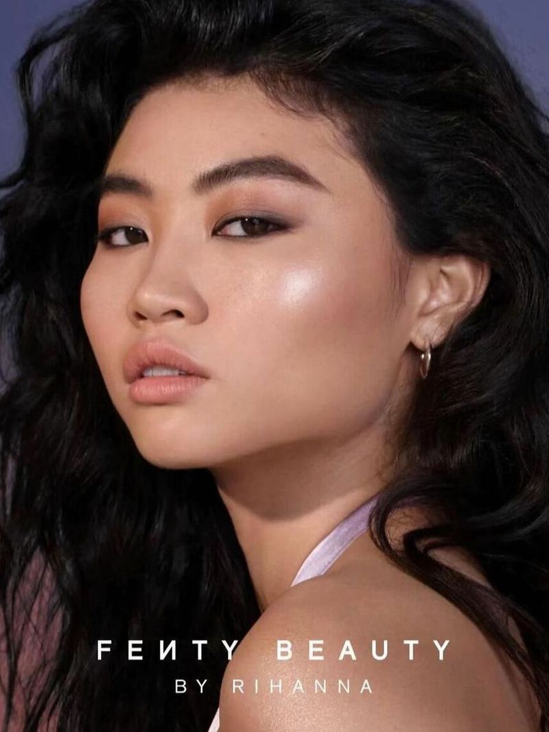 Close-up beauty shot of a young woman with dark hair and neutral makeup posing with her head tilted and looking at the camera.