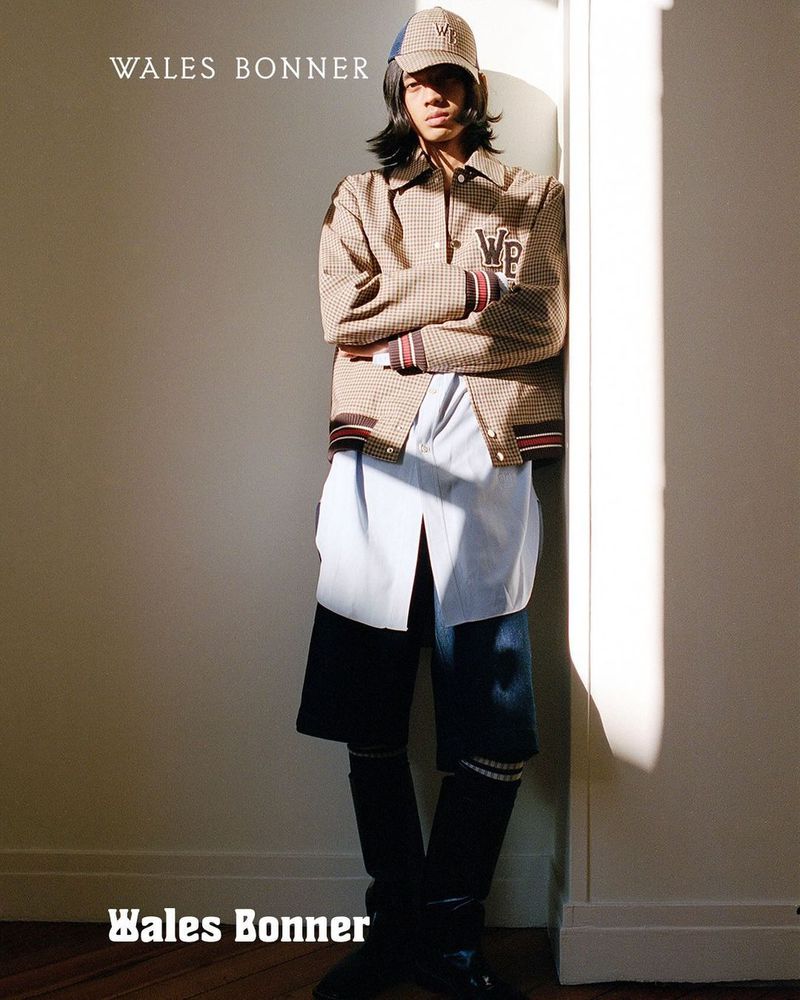 A young adult poses indoors wearing a Wales Bonner jacket, shirt, shorts, socks, boots, and hat in a fashion editorial shot.
