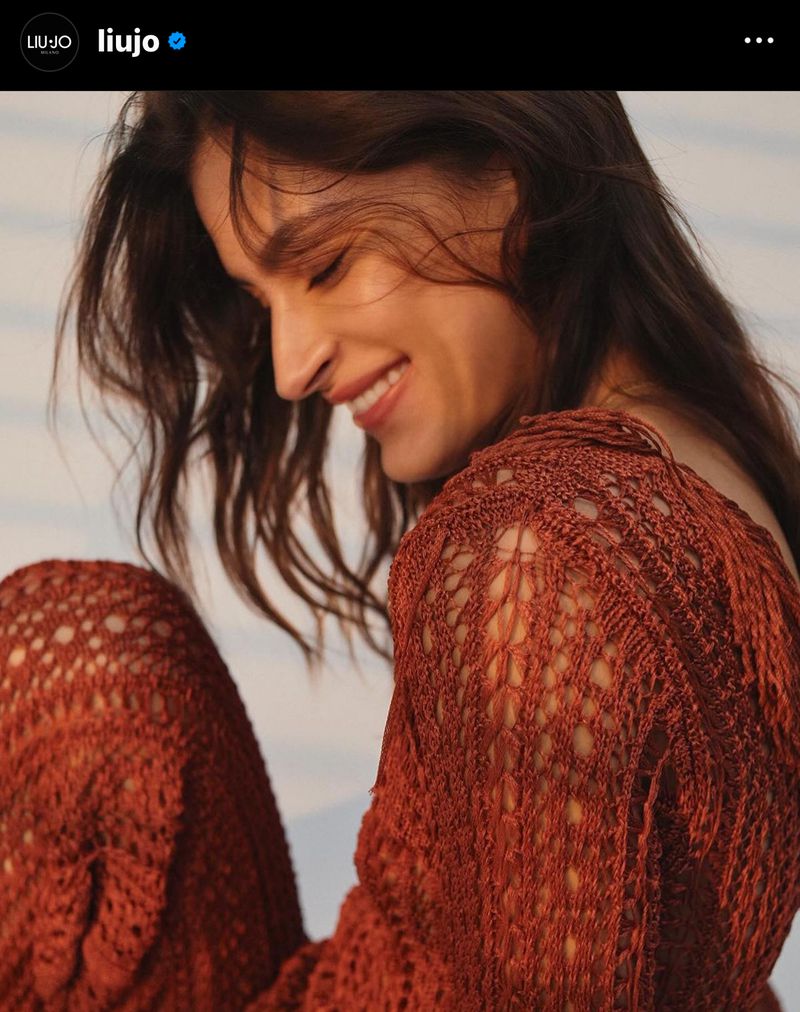 Smiling woman with brown hair wears a rust-colored knitted top, posing in front of a softly blurred, neutral background in a close-up shot.