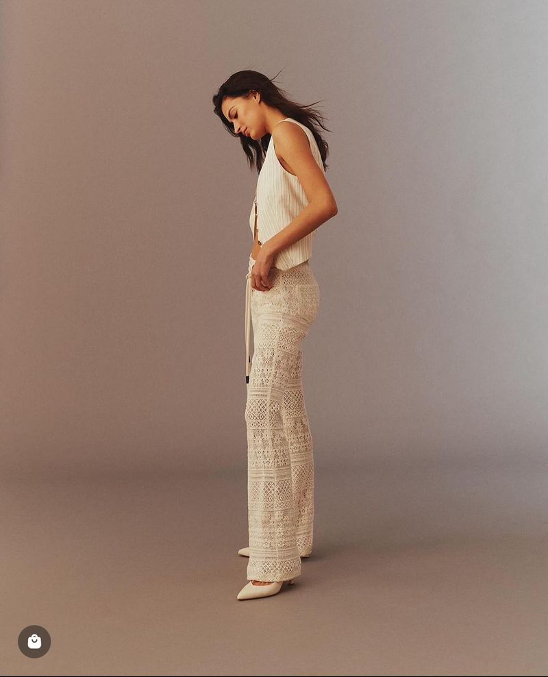 Elegant adult woman posing in a photo studio wearing a fashionable sleeveless top, crochet pants, and pointed heels in a side view.