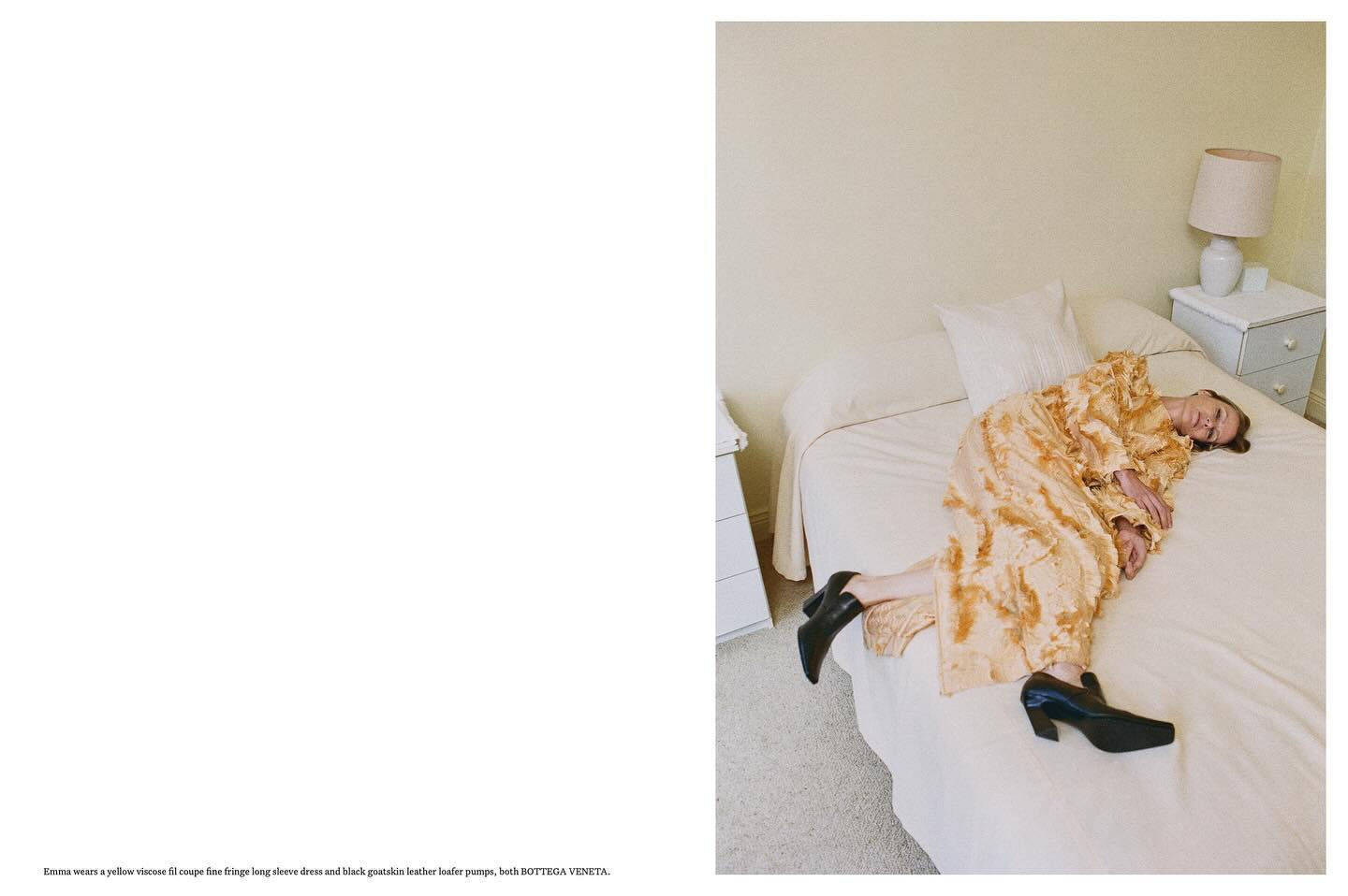 Adult woman in a viscose fringed dress and leather pumps rests on a white bed in an indoor environment with off-white walls.