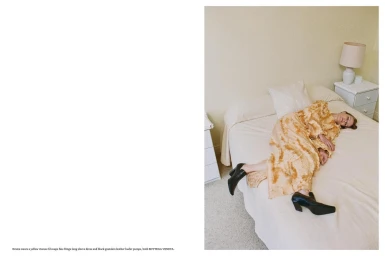 Model lying on a made bed wearing a textured orange dress and black leather shoes in a domestic bedroom.