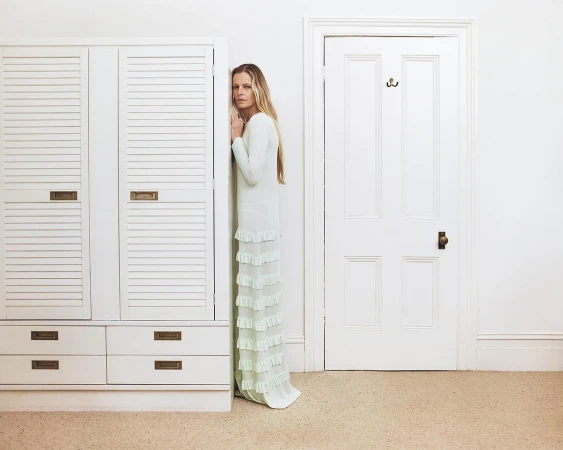 Adult female in a light green gown standing between a white wardrobe and white door inside a room with light walls and carpet.
