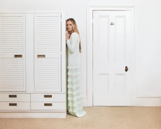 Woman with long blonde hair wearing a pale tiered dress leaning against a wall beside a white cabinet and door