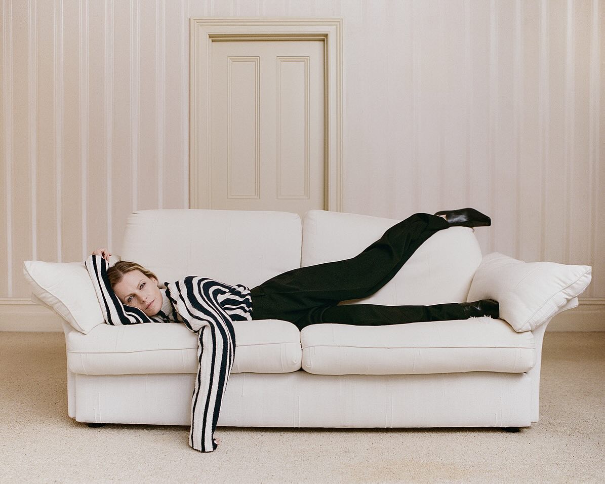 Woman in zebra top and black pants lounges on a white couch in a neutral room with a closed door, posing for fashion photography.