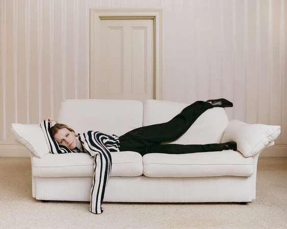 Woman in zebra top and black pants lounges on a white couch in a neutral room with a closed door, posing for fashion photography.