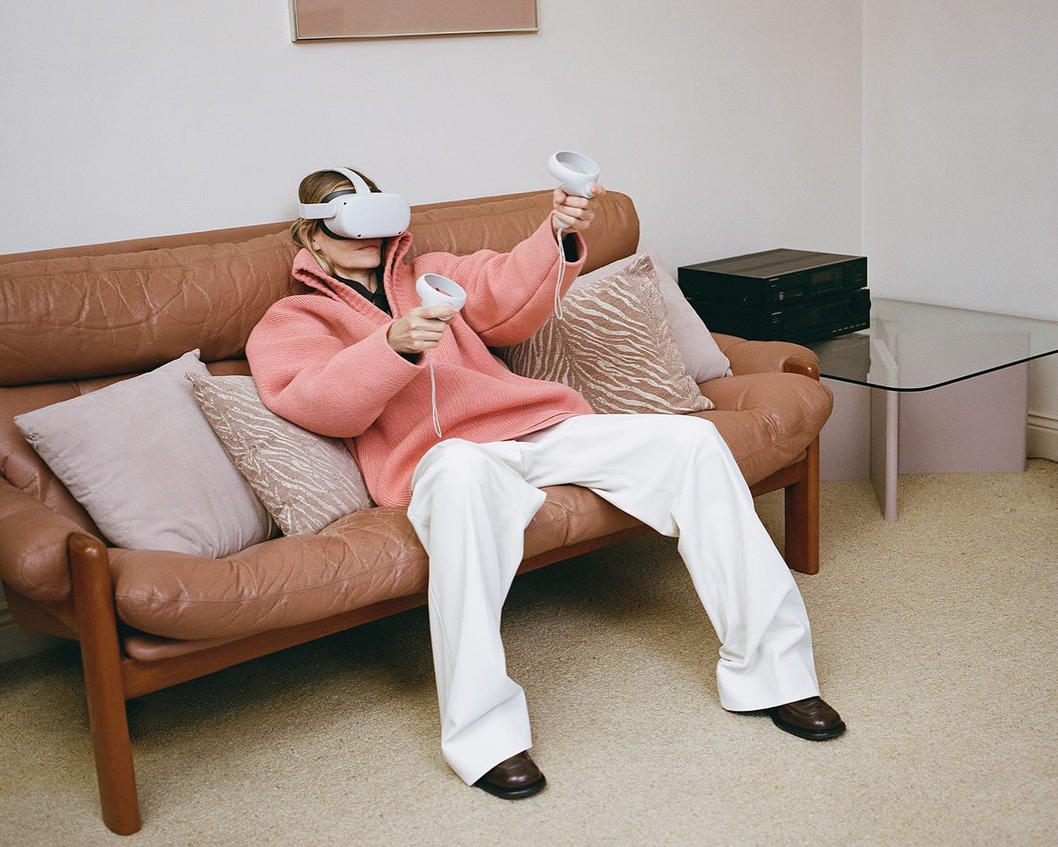 Woman wearing a virtual reality headset and holding controllers while sitting on a couch in a brightly lit living room.
