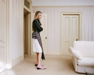Woman with long blonde hair in a black leather jacket and white skirt standing indoors near a white sofa