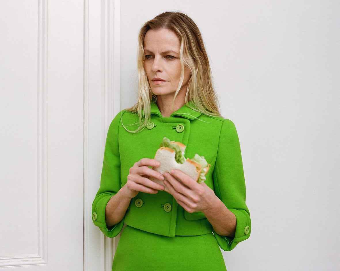 Adult woman in a green skirt suit eating a bitten sandwich while standing indoors near a white paneled wall, shot with a medium framing.
